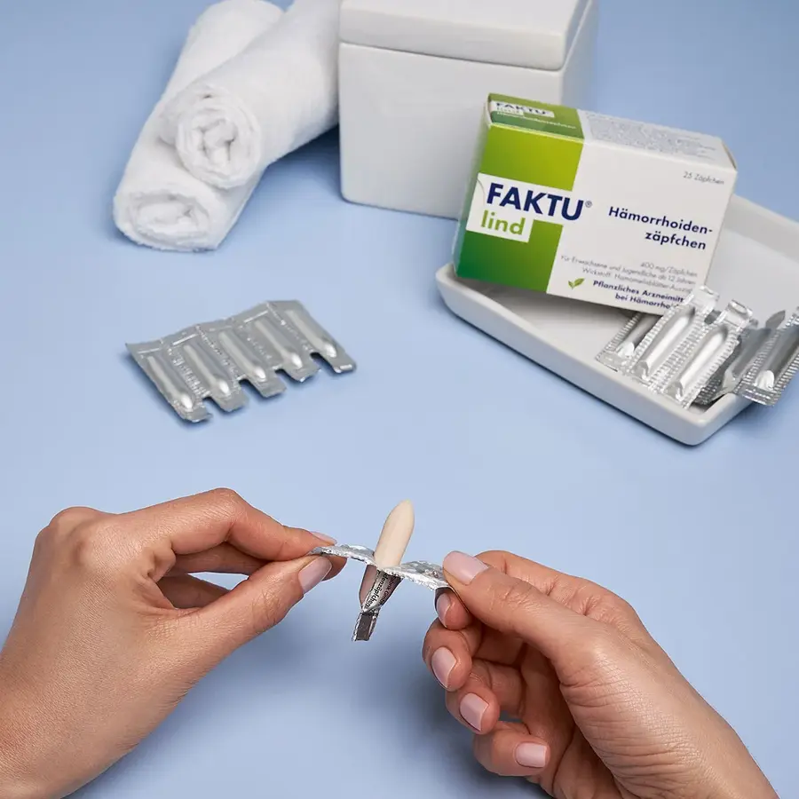 Hände öffnen ein FAKTU lind Zäpfchen aus der Blisterverpackung; im Hintergrund sind die Produktverpackung, weitere Zäpfchen in der Verpackung und gerollte weiße Handtücher.