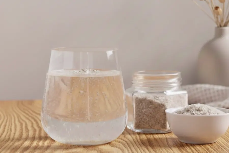 Glas mit sprudelndem Wasser auf einem Holztisch neben Schale und Glasdose mit Flohsamenschalen