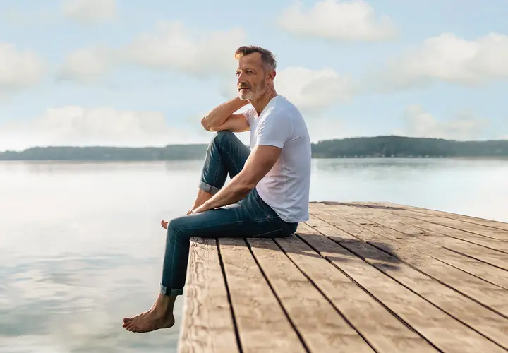 Älterer Mann sitzt am See und blickt nachdenklich in die Ferne.