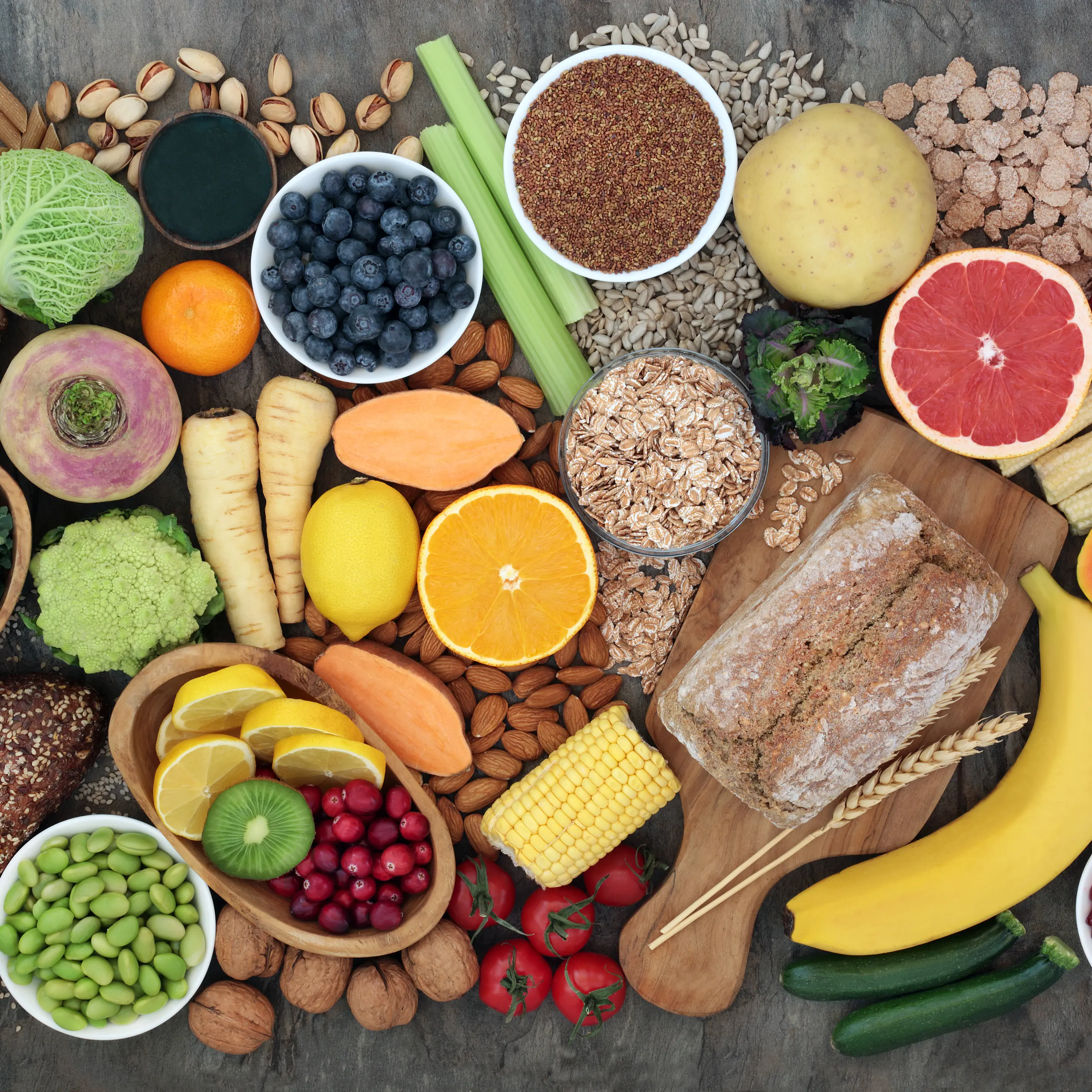 Verschiedene gesunde Lebensmittel wie Obst, Gemüse, Nüsse, Vollkornbrot, Haferflocken und Samen auf einem Tisch angerichtet