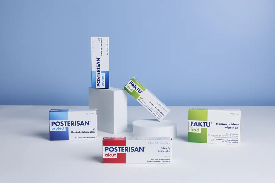 Arrangierte Produktdarstellung mit Posterisan akut, Posterisan protect Salbe und Zäpfchen sowie FAKTU lind Salbe und Zäpfchen auf einer weißen Fläche vor blauem Hintergrund.
