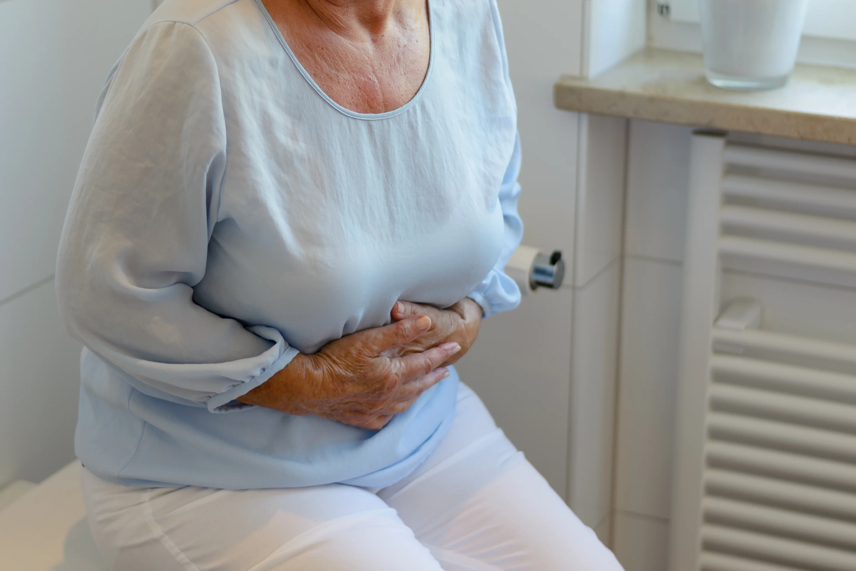 Ältere Person sitzt im Bad auf der geschlossenen Toilette und presst beide Hände auf den Unterbauch.