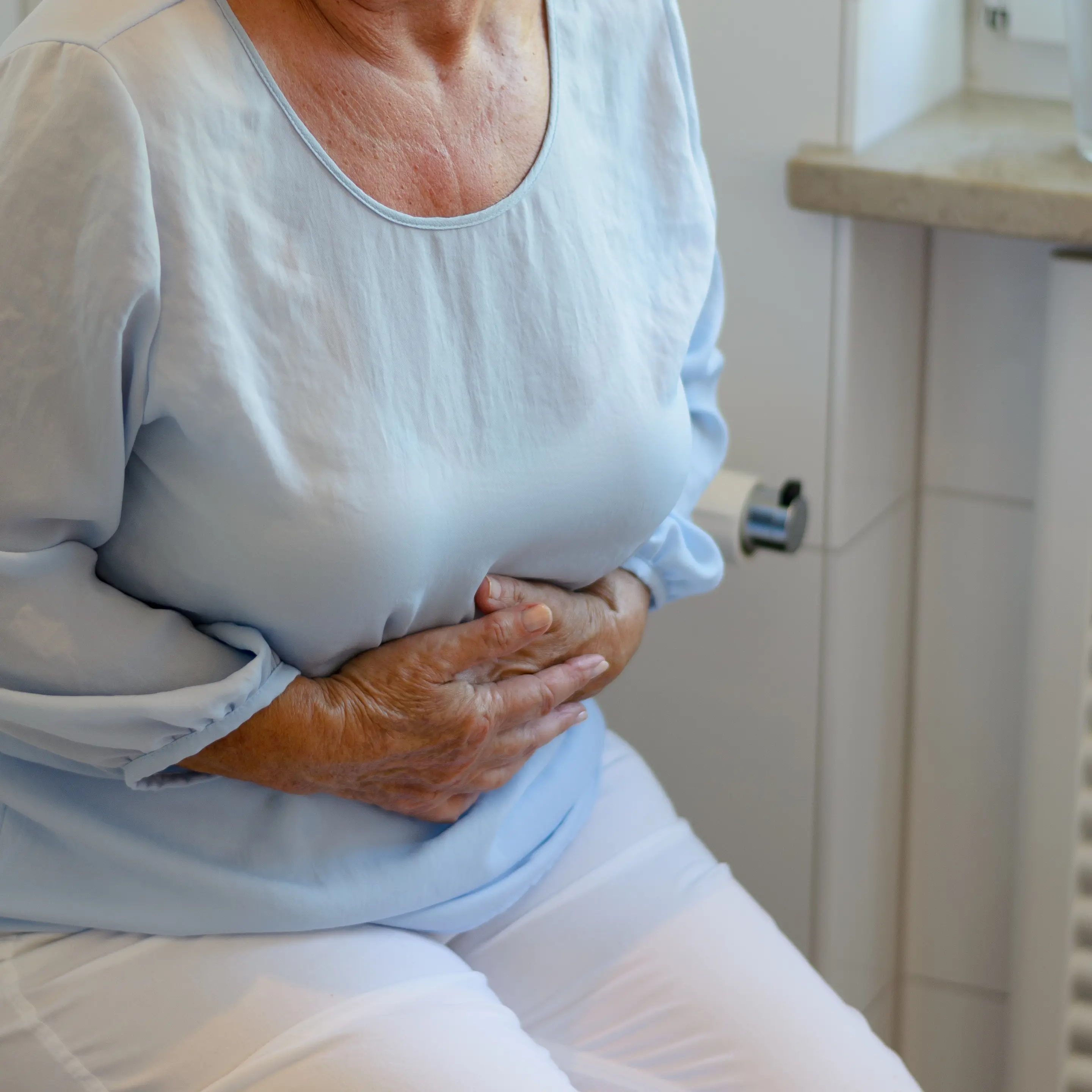 Ältere Person sitzt im Bad auf der geschlossenen Toilette und presst beide Hände auf den Unterbauch.