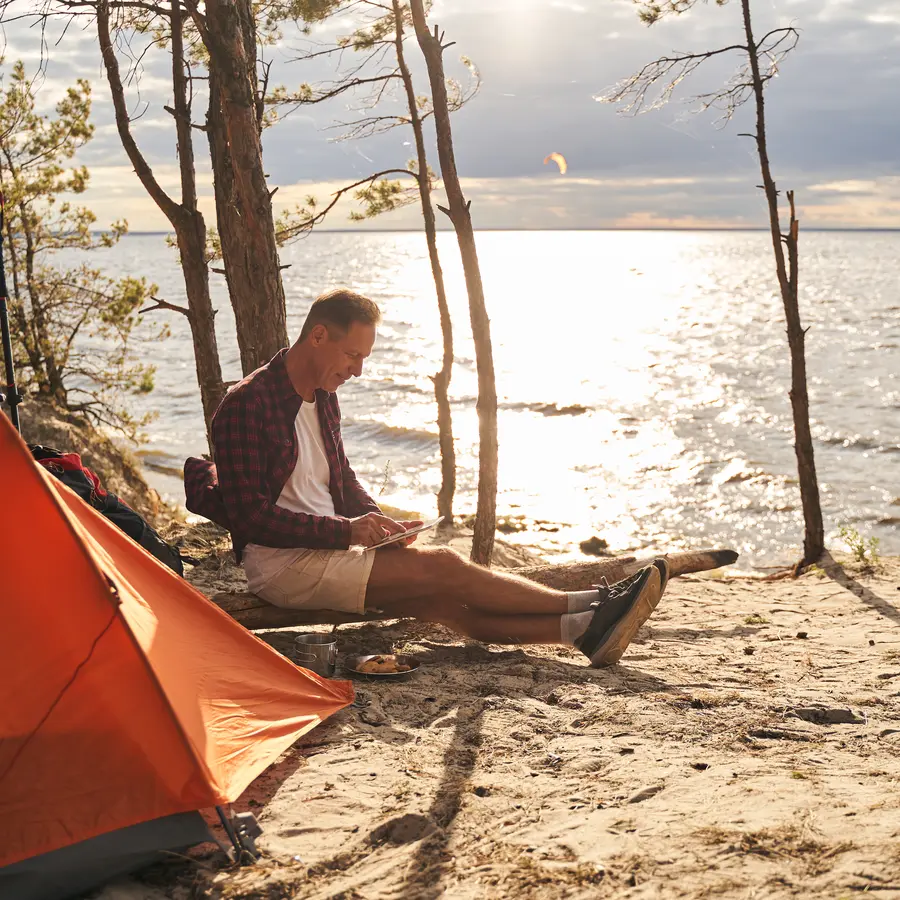 Mann sitzt beim Camping vor einem orangefarbenen Zelt am Ufer eines Sees und schaut auf ein Tablet.