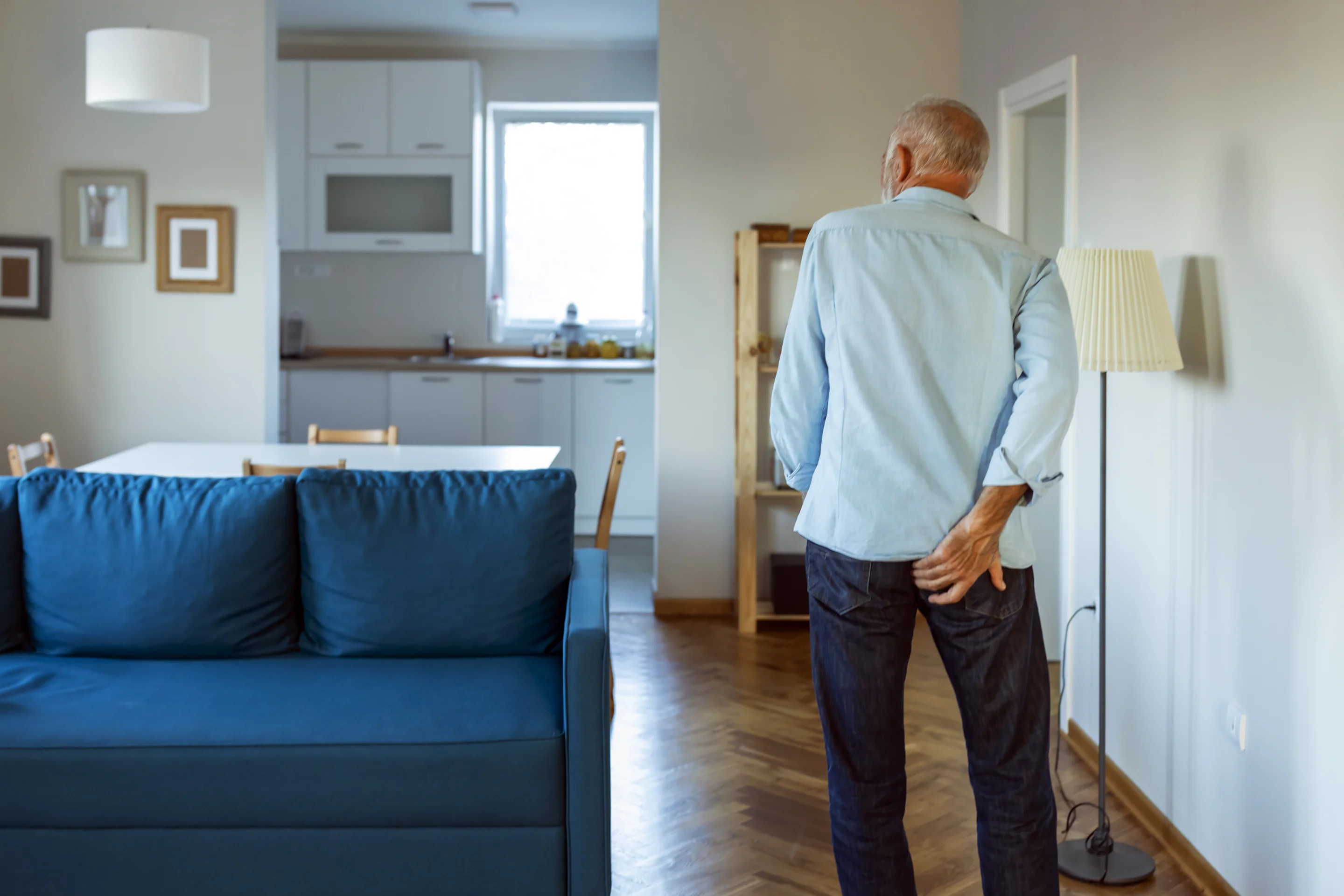 Ein älterer Mann steht in einem Wohnzimmer und fasst sich wegen Juckreiz am After ans Gesäß.