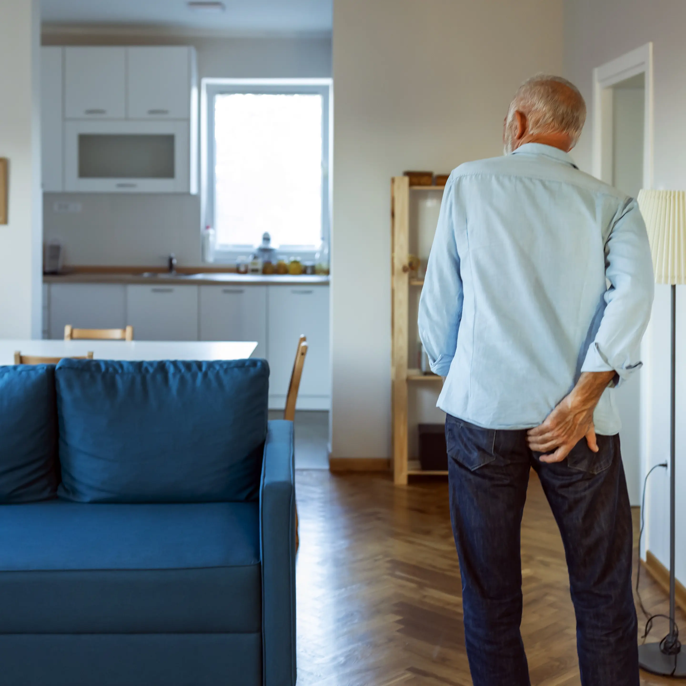 Ein älterer Mann steht in einem Wohnzimmer und fasst sich wegen Juckreiz am After ans Gesäß.