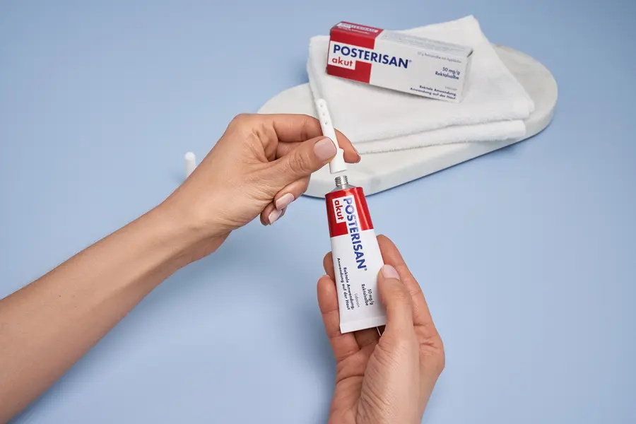 Person setzt Applikator auf eine Tube Posterisan akut Rektalsalbe; im Hintergrund die Verpackung auf einem gefalteten Handtuch.