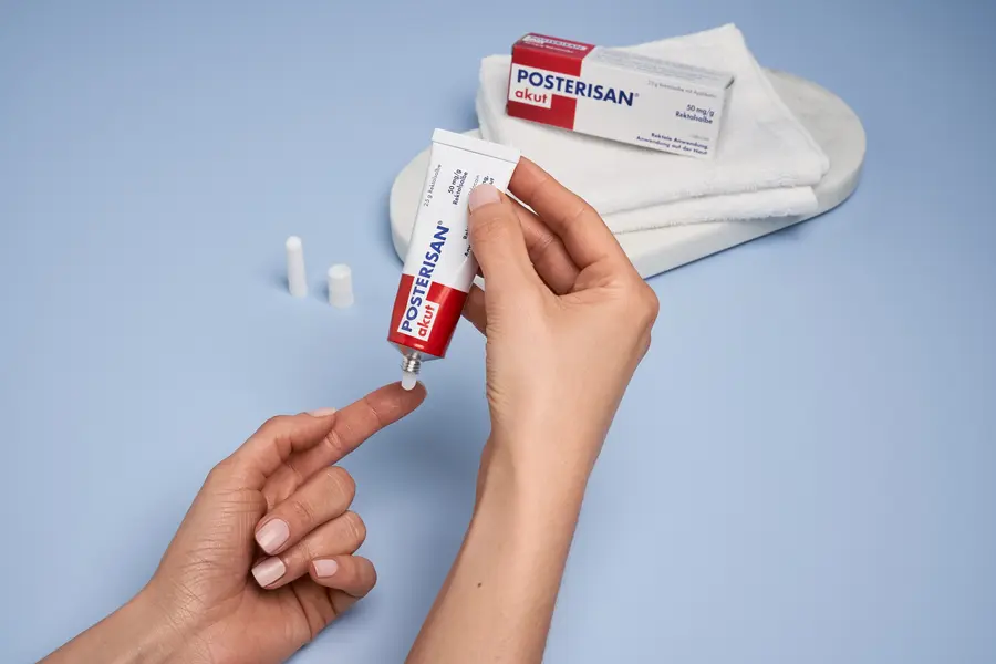 Hände halten eine Tube Posterisan akut Salbe und tragen die Hämorrhoiden-Salbe auf eine Fingerspitze auf.