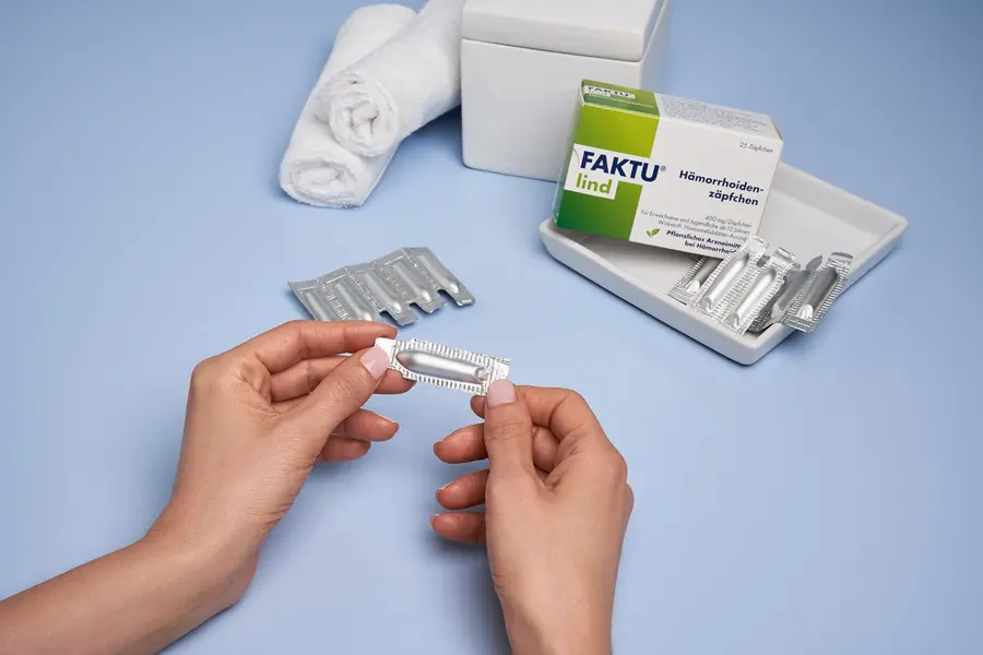 Person öffnet eine Blisterverpackung mit einem FAKTU lind Hämorrhoidenzäpfchen; im Hintergrund Packung, weitere Zäpfchen in einer Schale und gerollte Handtücher.