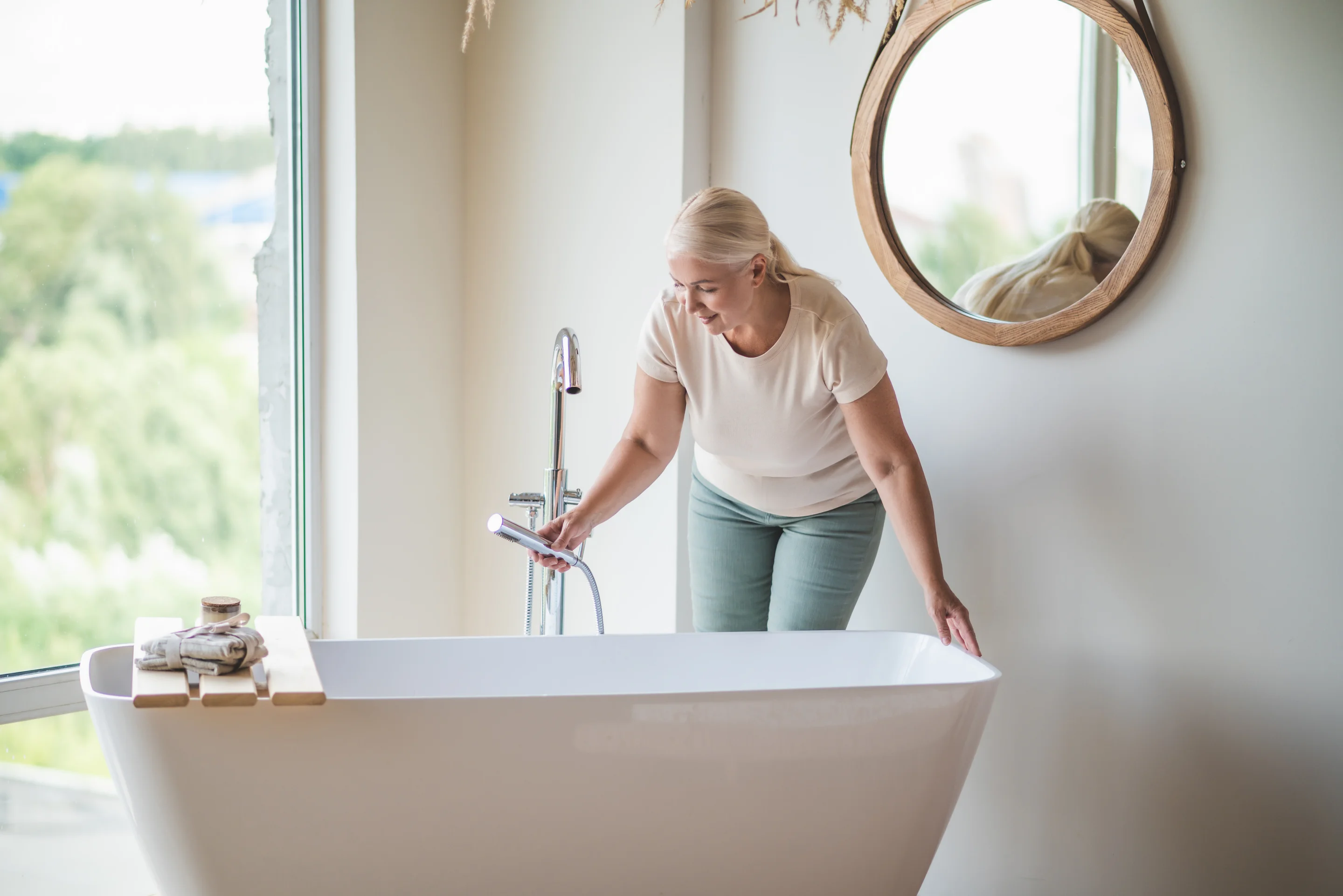Eine Frau beugt sich über eine frei stehende Badewanne in einem hellen Badezimmer und lässt Wasser einlaufen.