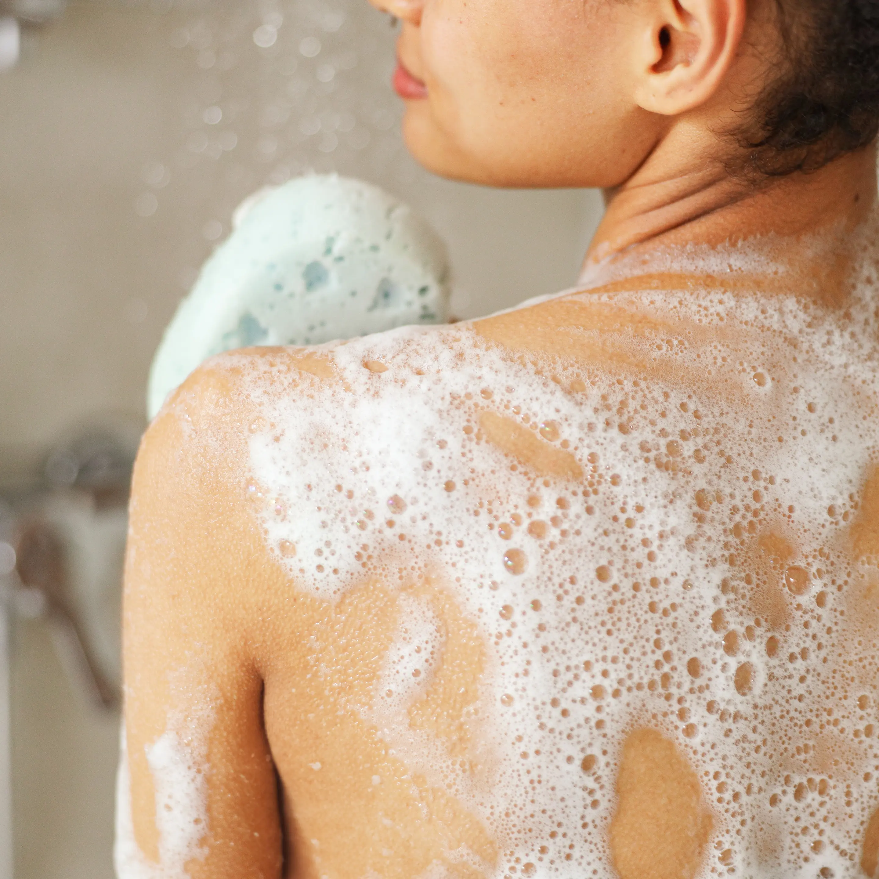 Frau steht unter der Dusche und seift sich den Rücken mit einem Schwamm ein.