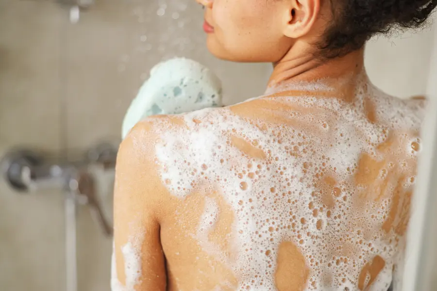 Frau steht unter der Dusche und seift sich den Rücken mit einem Schwamm ein.