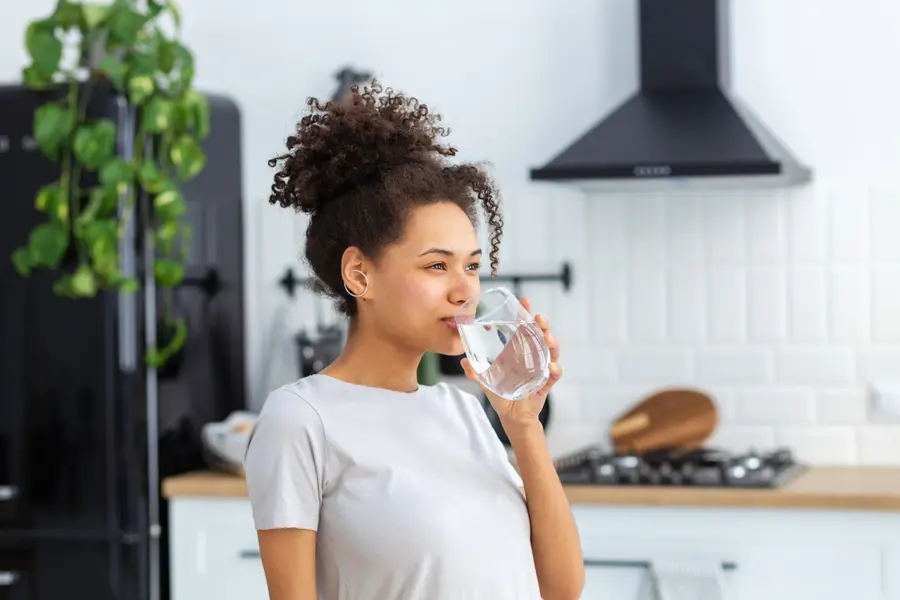Frau steht in einer hellen, modernen Küche und trinkt ein Glas Wasser.