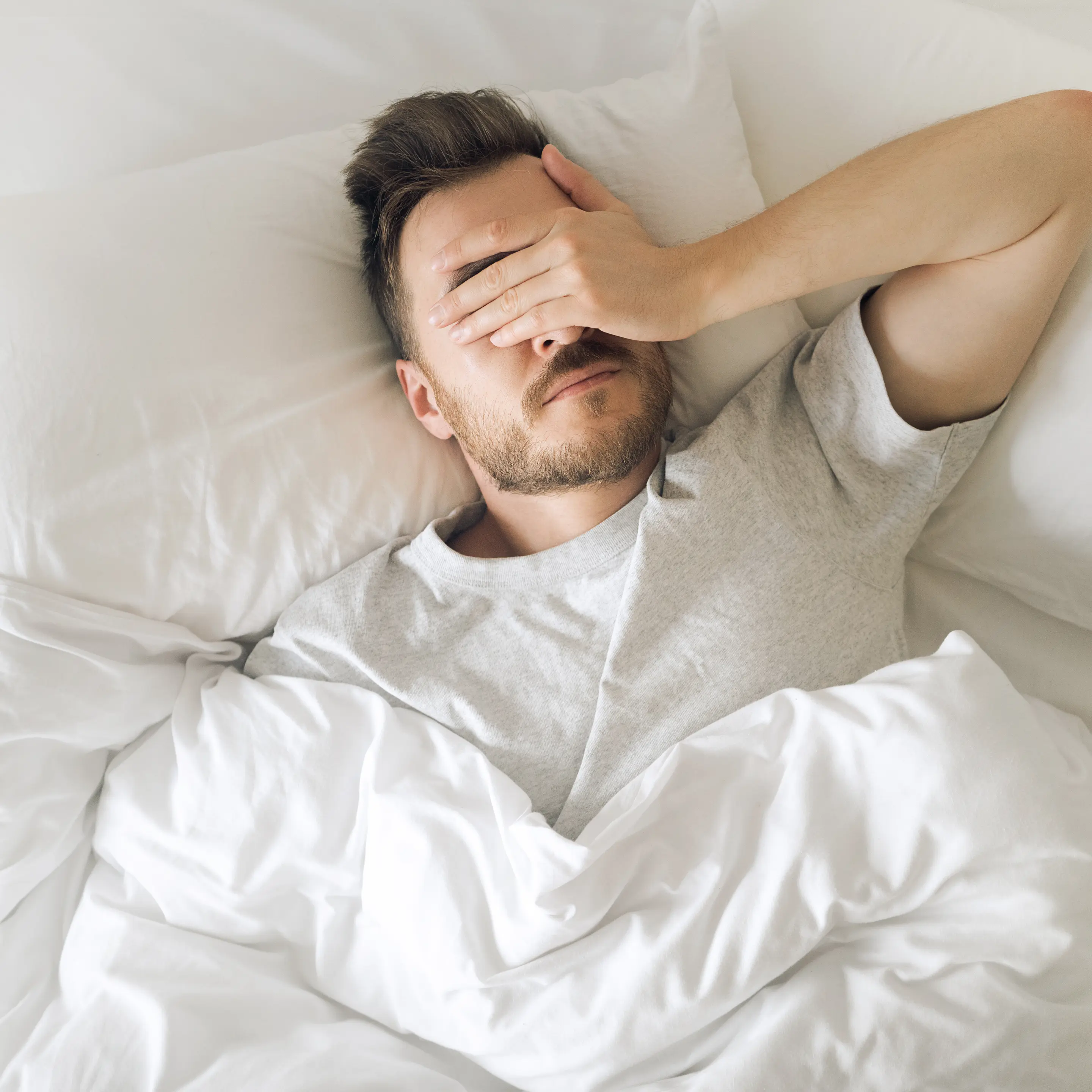 Ein Mann liegt im Bett und hält sich die Hand vors Gesicht.