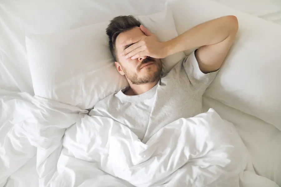 Ein Mann liegt im Bett und hält sich die Hand vors Gesicht.