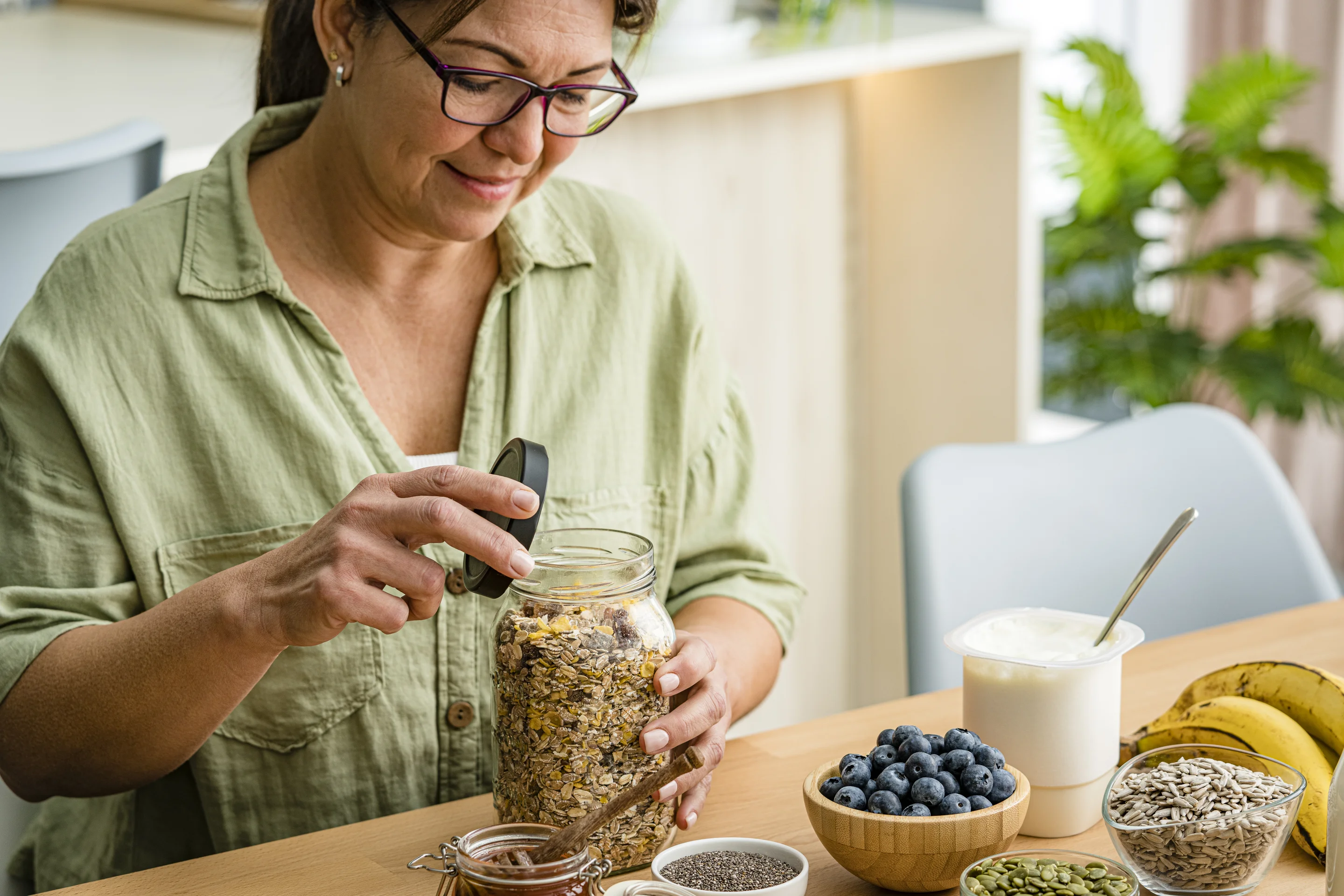 Person öffnet ein Glas mit Müsli an einem Tisch, auf dem Joghurt, Heidelbeeren, eine Banane sowie Schalen mit Kernen und Samen stehen.