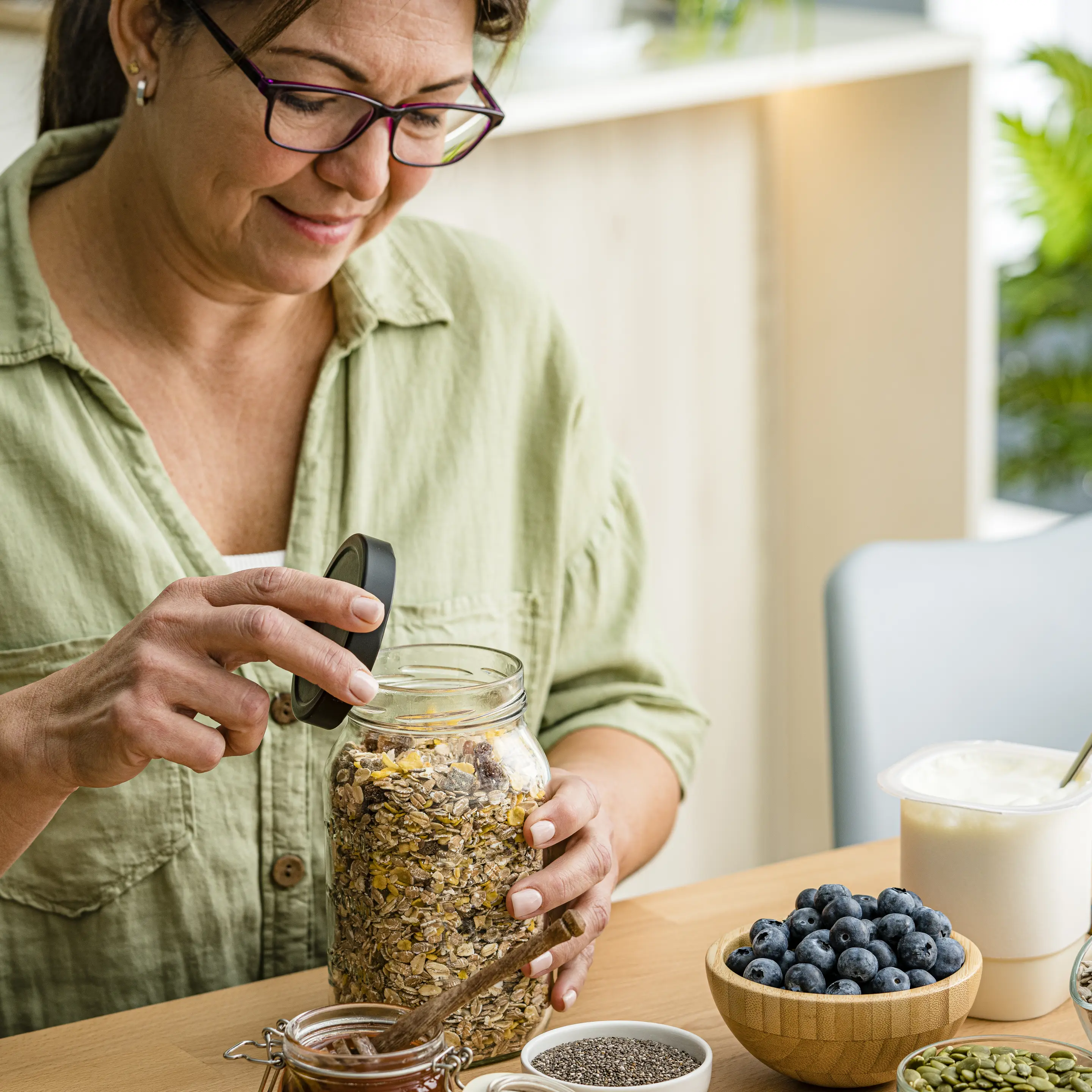 Person öffnet ein Glas mit Müsli an einem Tisch, auf dem Joghurt, Heidelbeeren, eine Banane sowie Schalen mit Kernen und Samen stehen.