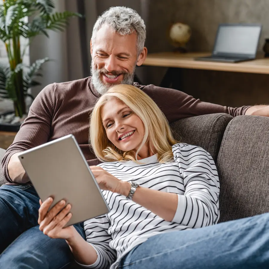 Lächelndes Paar entspannt gemeinsam auf dem Sofa und schaut auf ein Tablet.
