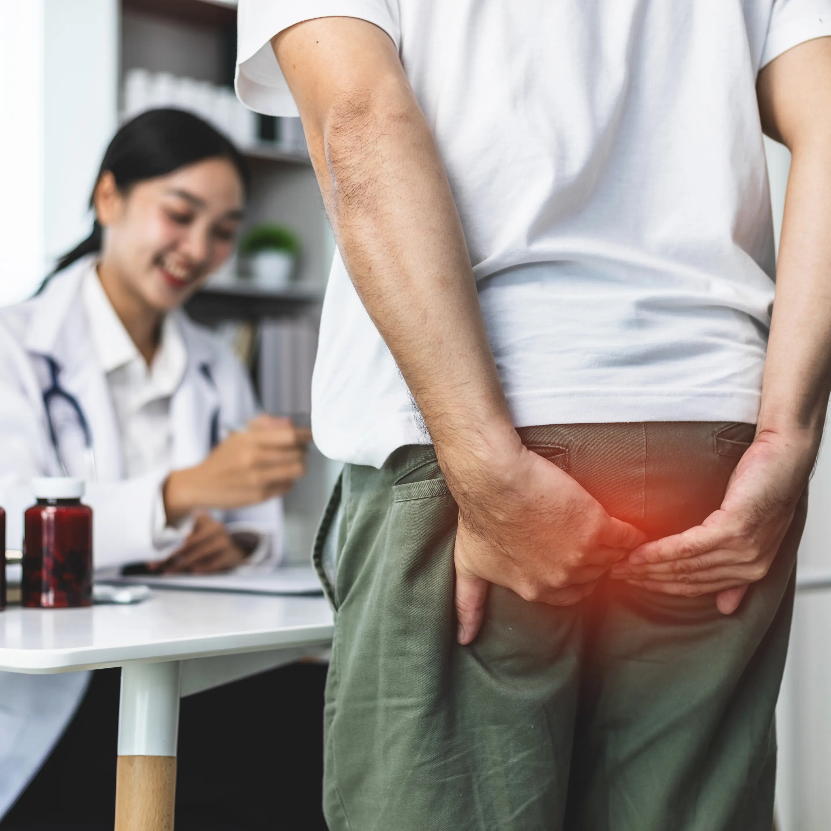 Patient steht in einer Praxis vor einer Ärztin und fasst sich an das Gesäß; eine farbige Markierung verdeutlicht Beschwerden im Analbereich.