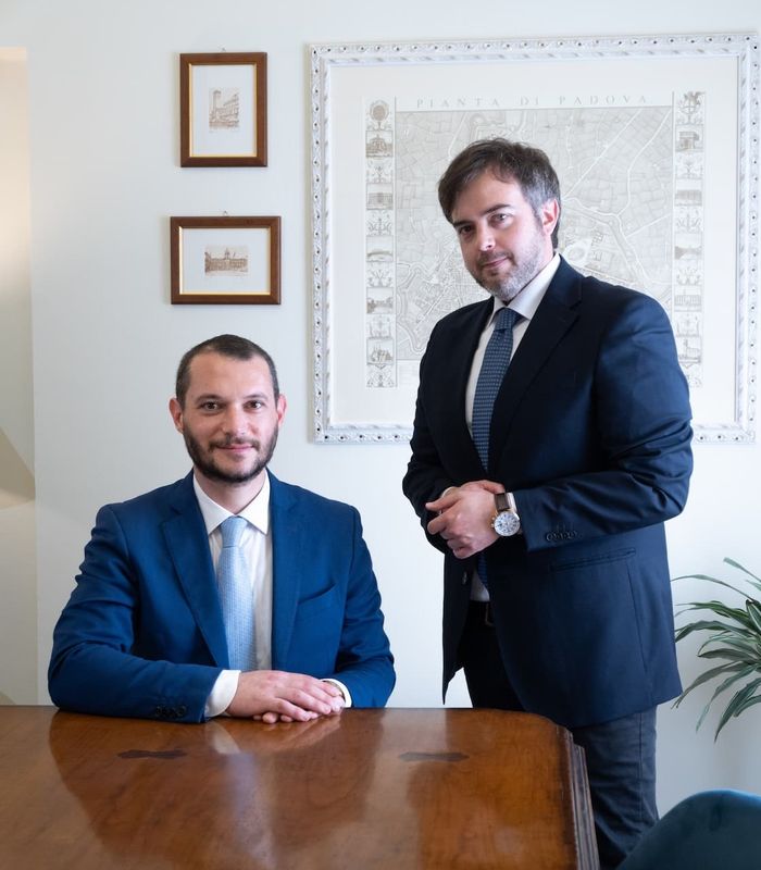 Two men in suits at a table, with framed pictures and a plant in the background, posing for a professional photo.