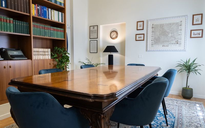 A cozy conference room with a wooden table, blue chairs, bookshelves, framed art, a clock, and plants, creating a professional atmosphere.