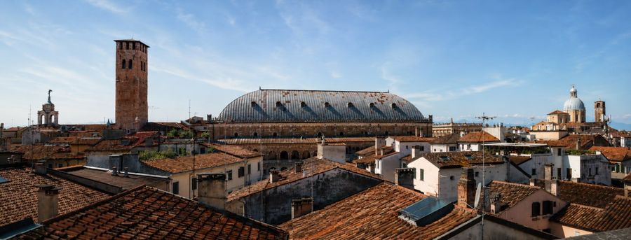 A panoramic view of a historic European city with red-tiled rooftops, a tall brick tower, and a large domed building under a clear blue sky.