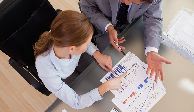 Two professionals discussing data at a desk, pointing at graphs and charts with pens, suggesting business analysis or financial review.
