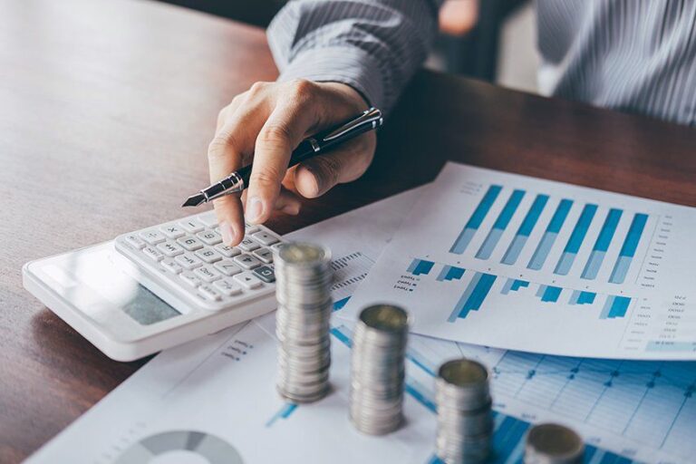 Person using a calculator with charts and stacked coins on a table, analyzing financial data.