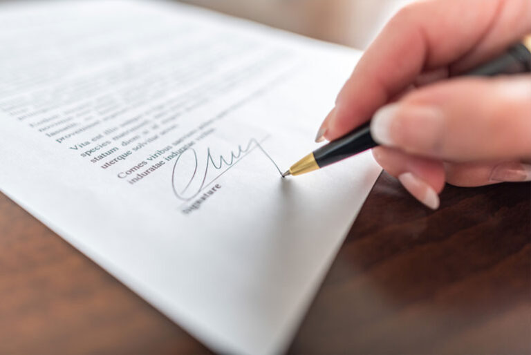 A hand signing a document with a black pen on a wooden table. The signature is at the bottom of a typed page.