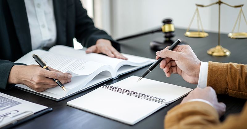Two people reviewing legal documents at a desk with a gavel and scales of justice in the background.
