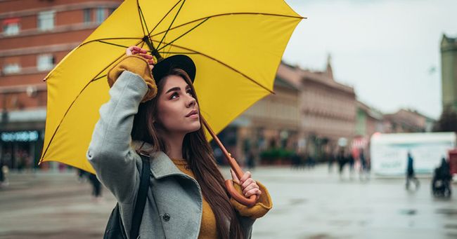 Woman in a gray coat holding a yellow umbrella, standing on a city street with blurred buildings and pedestrians in the background.