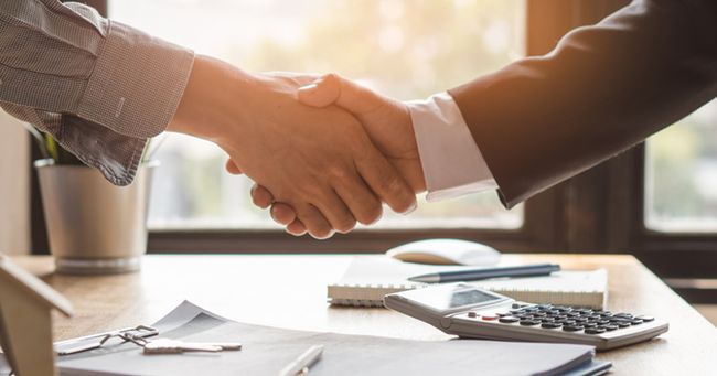 Two people in suits shaking hands over a desk with papers, a calculator, and a window in the background.
