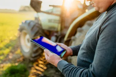 Ein Landwirt steht neben einem Traktor auf einem Feld und hält ein Tablet in den Händen bei Sonnenuntergang.