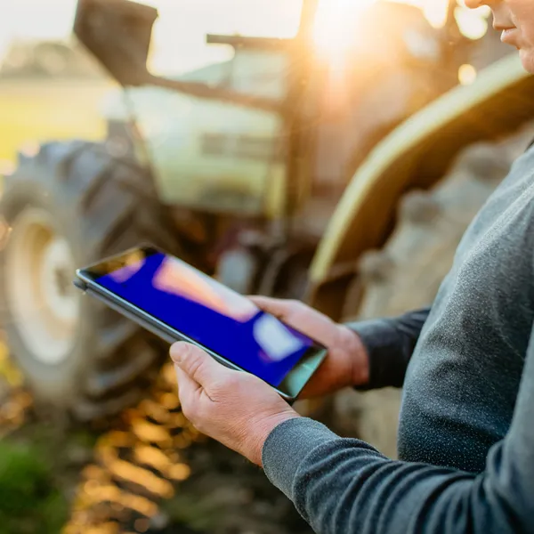 Ein Landwirt steht neben einem Traktor auf einem Feld und hält ein Tablet in den Händen bei Sonnenuntergang.