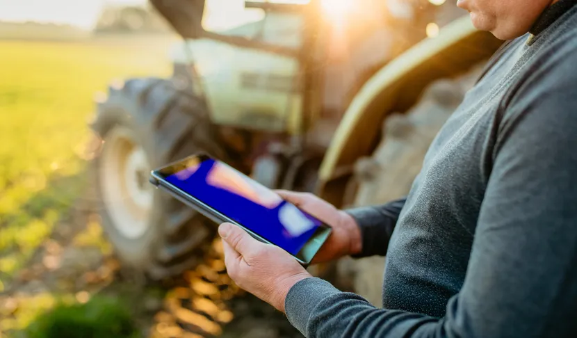 Ein Landwirt steht neben einem Traktor auf einem Feld und hält ein Tablet in den Händen bei Sonnenuntergang.