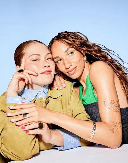 Two women pose closely against a blue background; one with braided hair and tattoos, the other with slicked-back hair and vibrant nail polish.