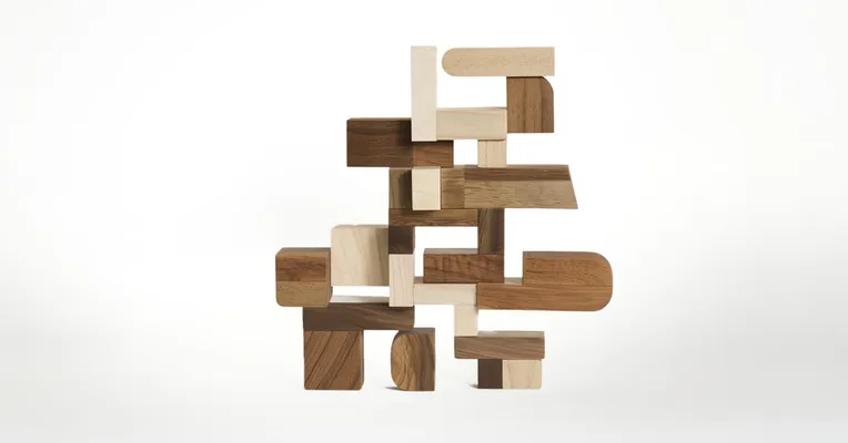 An abstract sculpture of stacked, mismatched wooden offcuts in varying tones and shapes, balanced into a freeform tower against a plain white background