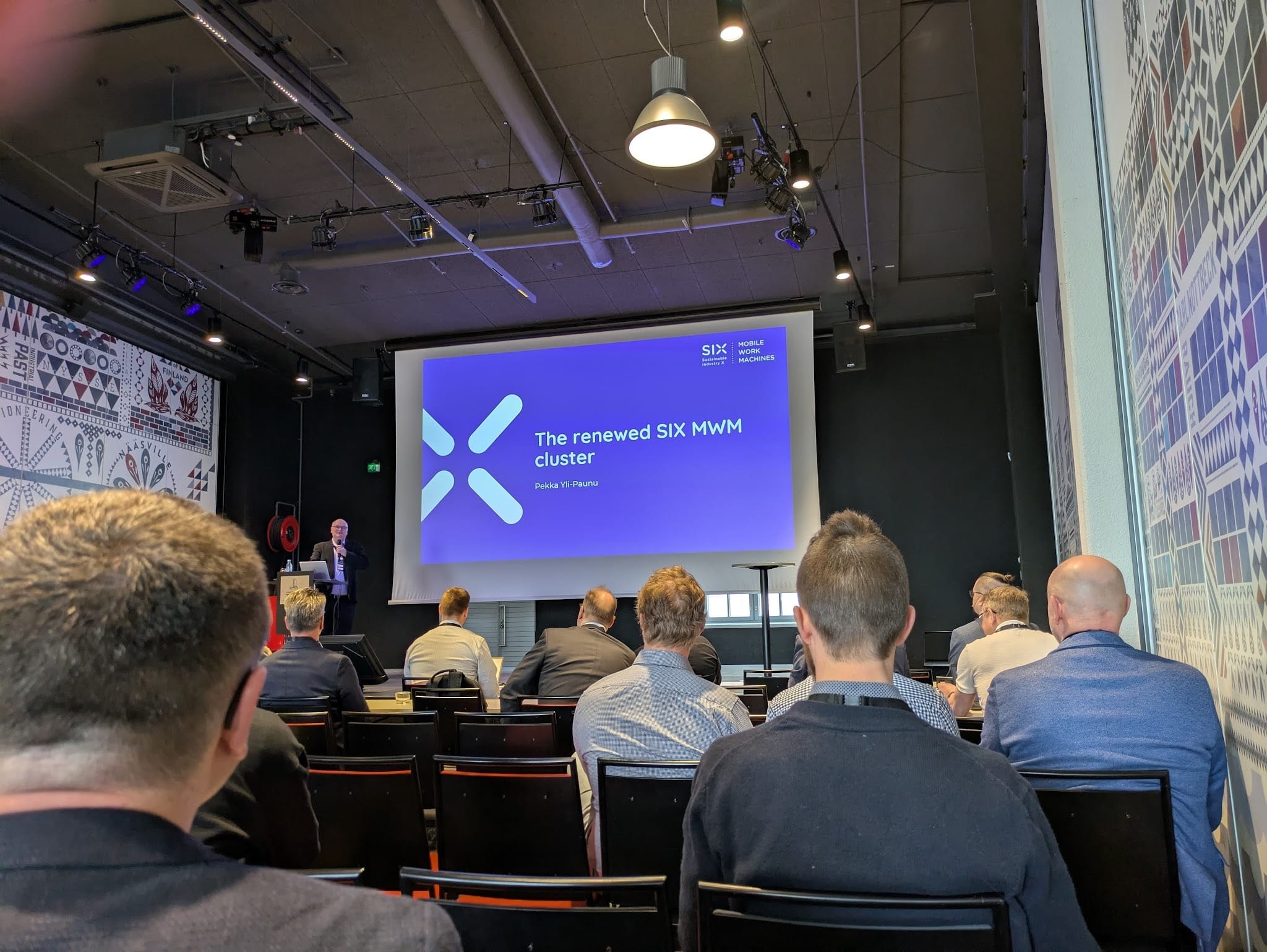 Seminar audience seated with their backs to the camera, watching a presentation titled 'The renewed SIX MWM cluster' by Pekka Yli-Paunu on a large screen