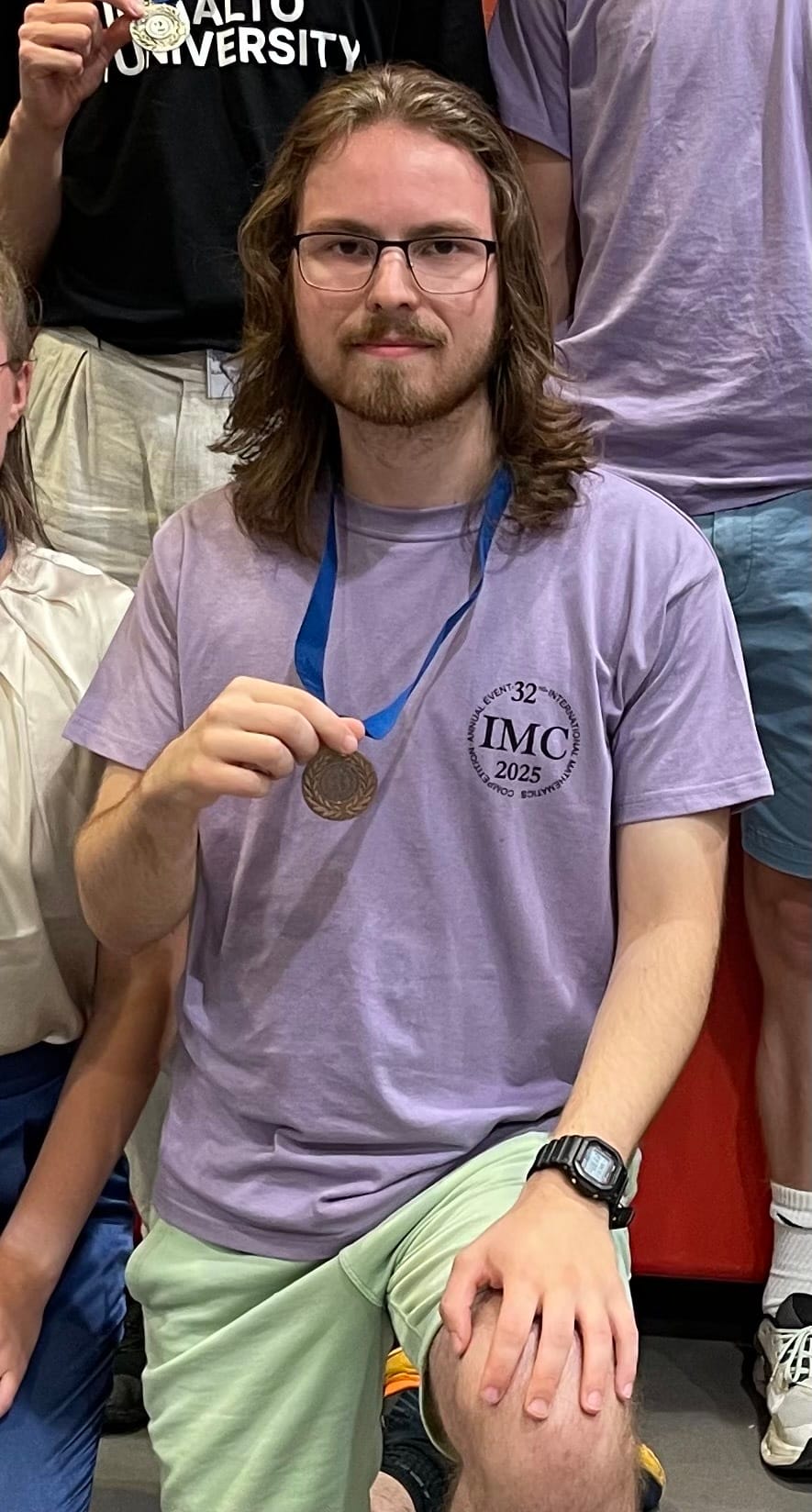 Sampo Siitonen kneeling and holding up his bronze medal, wearing a lilac '32nd International Mathematics Competition IMC 2025' T-shirt, with a teammate from Aalto University behind him holding up a gold medal