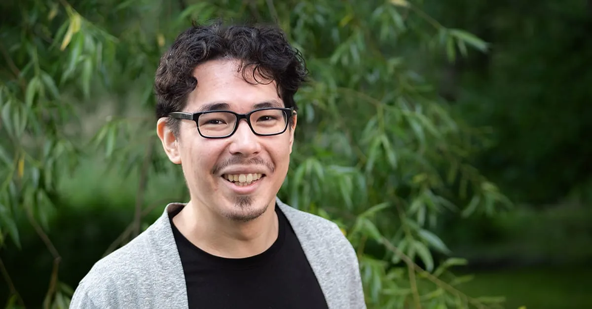 Portrait of Mitja smiling outdoors in front of green willow foliage, wearing a grey cardigan over a black top