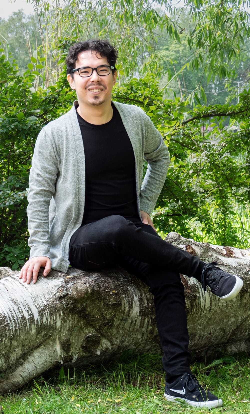 Mitja sitting on a birch log in front of green willow foliage, wearing a grey cardigan over a black top