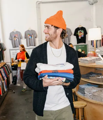 Un dependiente con gorro naranja cruza la tienda con una pila de camisetas dobladas