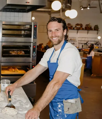 A baker in a blue apron at a floury worktable, the ovens behind him