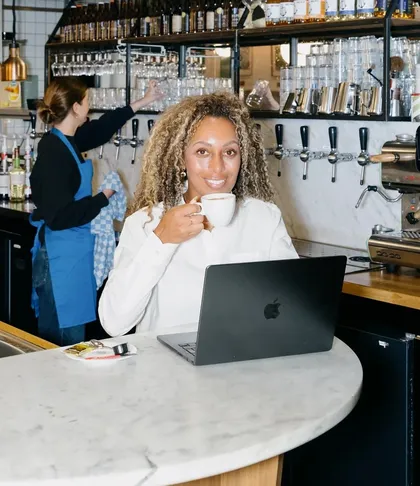 Una propietaria de restaurante en la barra con un café y su portátil, con una compañera trabajando detrás