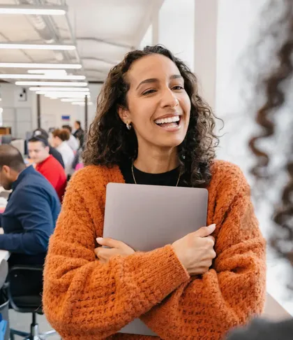 A woman in an orange jumper laughing, her laptop held to her chest, in a busy office