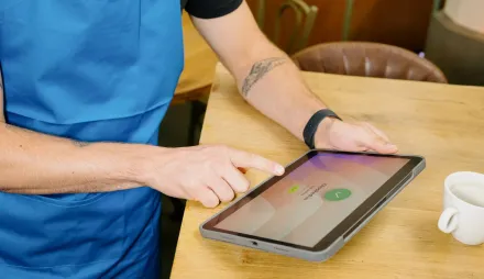 Ein Mitarbeiter mit blauer Schürze stempelt an einem Cafétisch auf einem Tablet ein