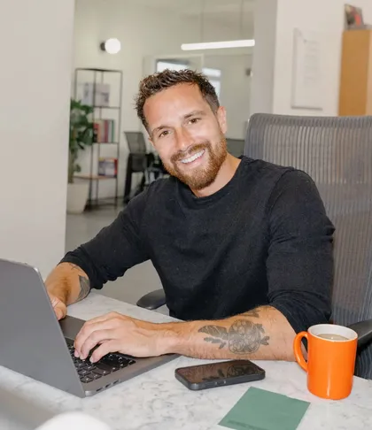 Un hombre en su escritorio levanta la vista del portátil sonriendo, con una taza naranja al lado