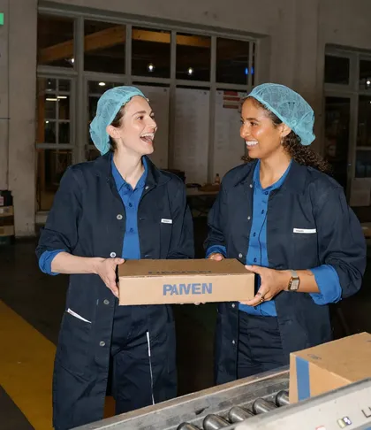 Zwei Produktionsmitarbeiterinnen mit Haarnetz und blauer Arbeitskleidung tragen lachend gemeinsam einen Karton