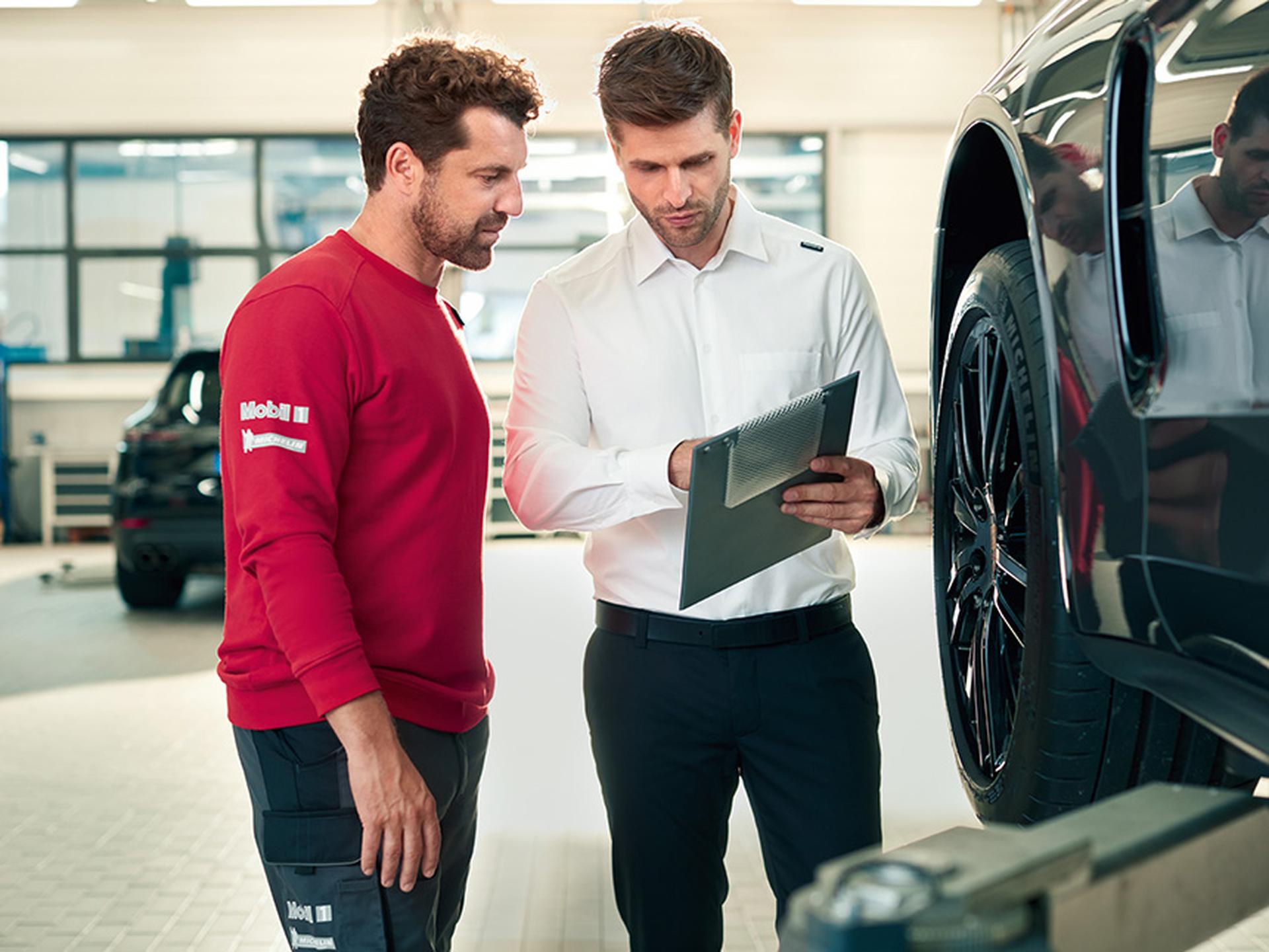 Zwei Porsche Mitarbeiter besprechen sich an der Frontachse eines Fahrzeugs. Einer hält ein Klemmbrett in Händen.