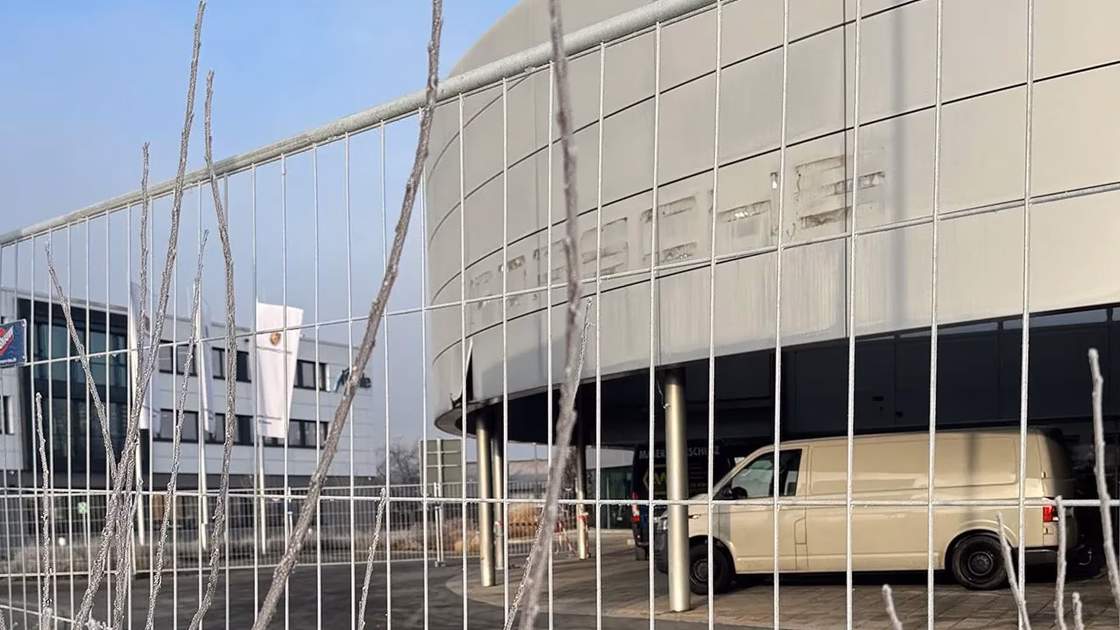 Porsche Zentrum mit abgerundeter Fassade und parkendem weißen Van im Vordergrund, davor ein Bauzaun mit vertikalen Stäben.